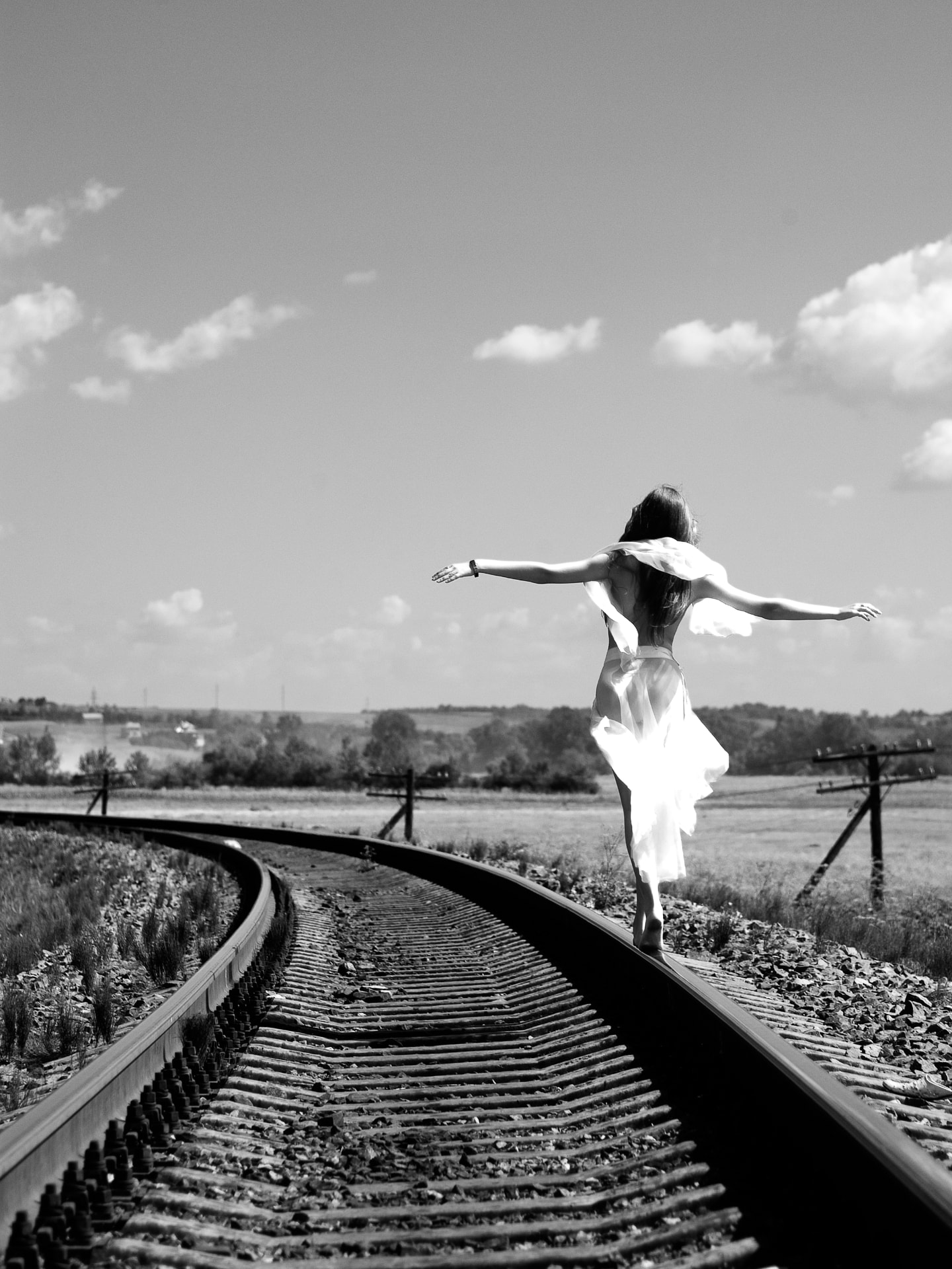 A woman in a flowing white dress balancing on railroad tracks, arms outstretched, beneath an open sky